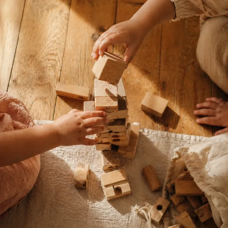 Two children's hands building a tower of natural wooden blocks on a warm oak floor